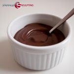 A bowl of smooth chocolate pudding with a spoon resting in it, branded with 'Strange Sculpting