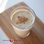 A glass of Cinnamon Toast Protein Smoothie on a wooden cutting board, with a sprinkle of cinnamon on top
